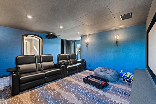 carpeted cinema room featuring arched walkways, visible vents, and a textured wall