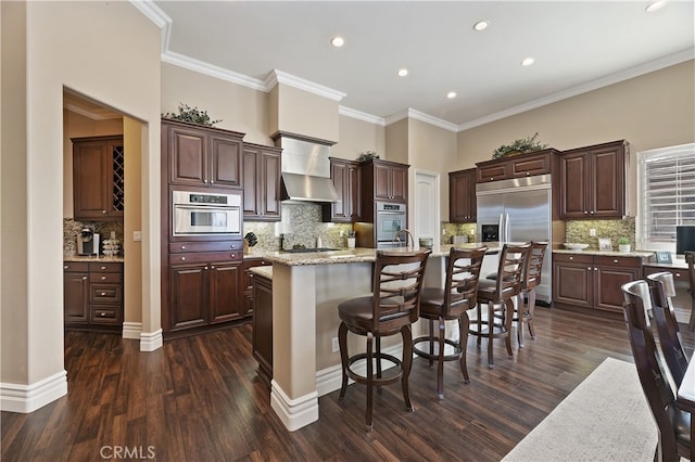 kitchen featuring an island with sink, wall chimney range hood, appliances with stainless steel finishes, and dark brown cabinets