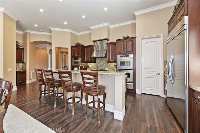 kitchen featuring a center island with sink, arched walkways, wall chimney exhaust hood, light stone countertops, and stainless steel appliances