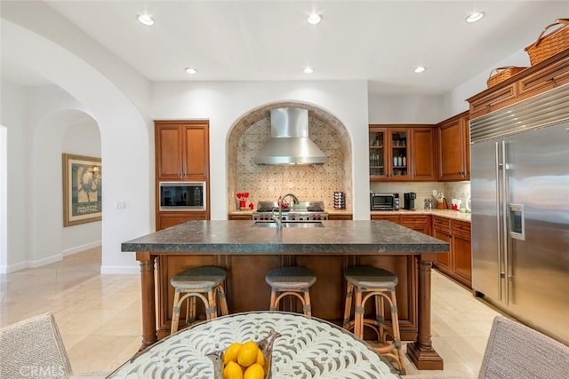 kitchen featuring built in appliances, wall chimney exhaust hood, a breakfast bar, and arched walkways