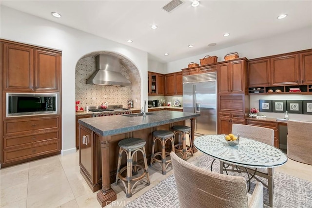 kitchen with built in appliances, a breakfast bar, decorative backsplash, wall chimney exhaust hood, and a center island with sink