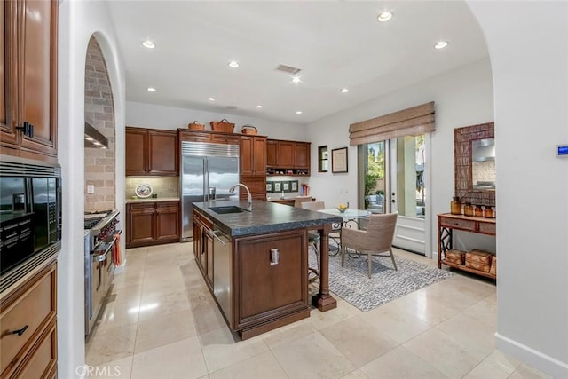 kitchen featuring a center island with sink, decorative backsplash, dark countertops, a kitchen breakfast bar, and built in appliances