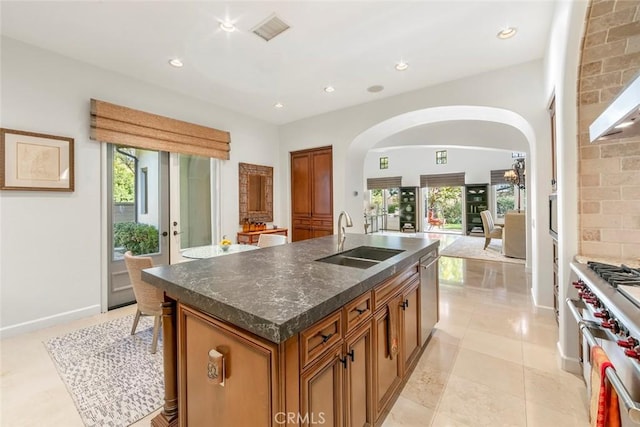 kitchen with arched walkways, a sink, appliances with stainless steel finishes, brown cabinets, and a center island with sink