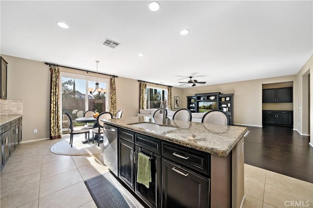 kitchen featuring light stone counters, a kitchen island with sink, a sink, stainless steel dishwasher, and pendant lighting