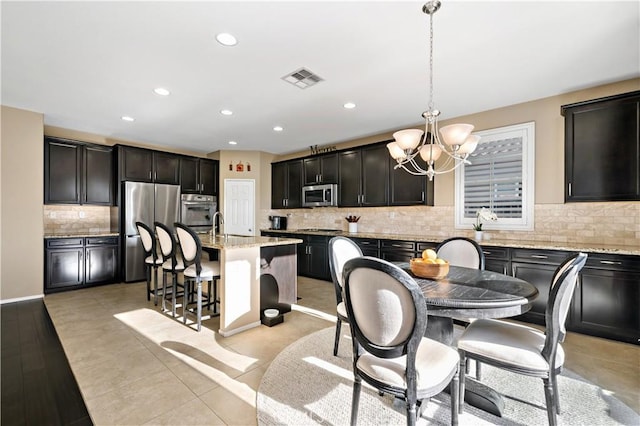 kitchen with a kitchen island with sink, a notable chandelier, stainless steel appliances, light stone countertops, and pendant lighting