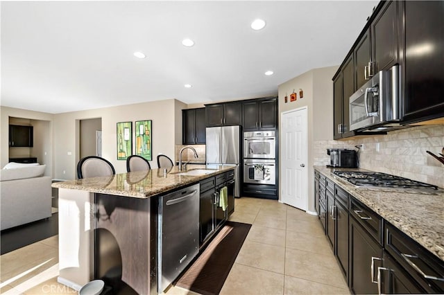 kitchen featuring stainless steel appliances, a kitchen island with sink, a sink, light tile patterned flooring, and a kitchen bar