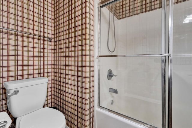 bathroom featuring toilet, bath / shower combo with glass door, and tile walls