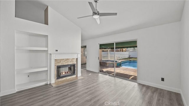 unfurnished living room with built in shelves, a fireplace, wood finished floors, a ceiling fan, and baseboards