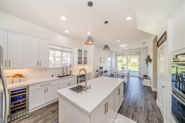 kitchen with hanging light fixtures, glass insert cabinets, a kitchen island with sink, white cabinets, and beverage cooler