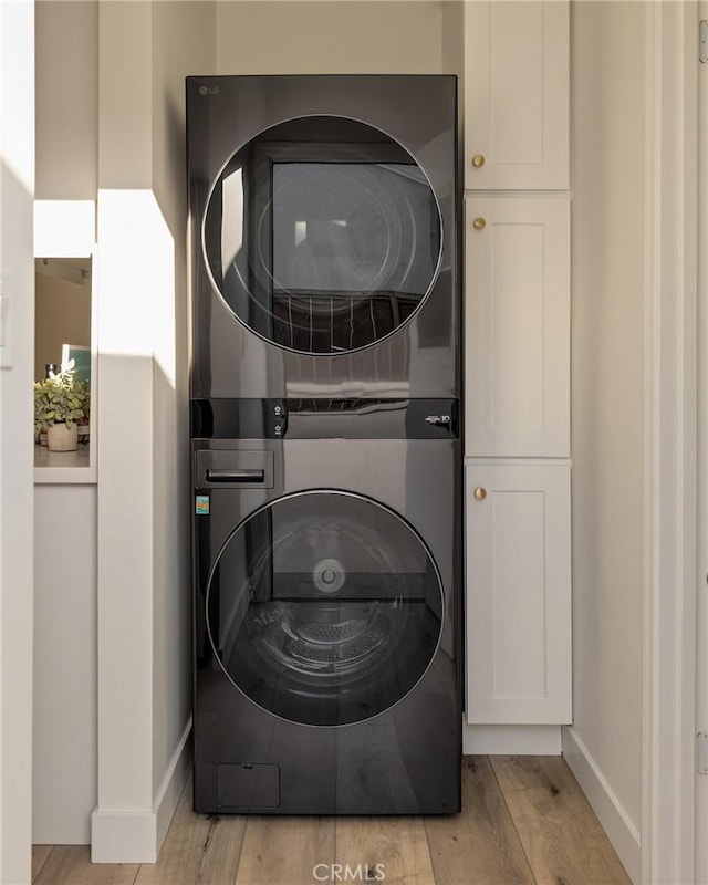 clothes washing area featuring light wood-style floors, stacked washer and clothes dryer, cabinet space, and baseboards