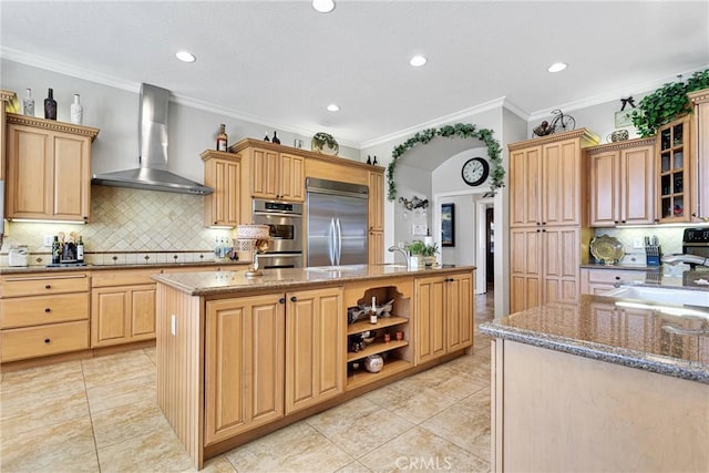 kitchen featuring a center island with sink, wall chimney exhaust hood, glass insert cabinets, appliances with stainless steel finishes, and light stone countertops