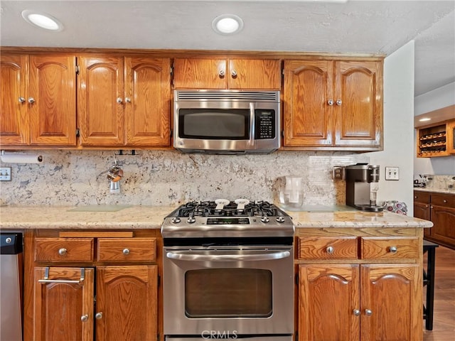 kitchen with brown cabinets, backsplash, appliances with stainless steel finishes, light wood-style floors, and light stone countertops