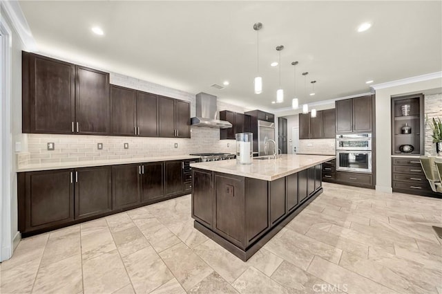 kitchen featuring an island with sink, wall chimney range hood, dark brown cabinetry, and decorative light fixtures