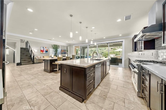 kitchen featuring a sink, exhaust hood, appliances with stainless steel finishes, an island with sink, and pendant lighting