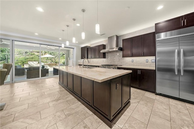 kitchen with decorative light fixtures, light countertops, a kitchen island with sink, stainless steel built in refrigerator, and wall chimney exhaust hood