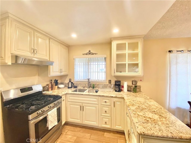 kitchen featuring gas range, glass insert cabinets, light stone counters, under cabinet range hood, and a sink