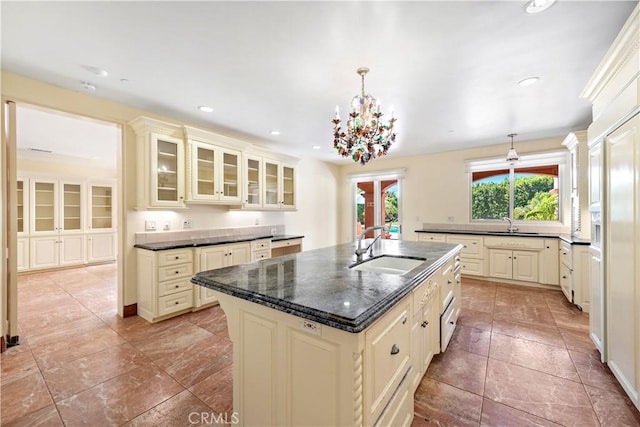 kitchen with a sink, hanging light fixtures, cream cabinetry, an island with sink, and glass insert cabinets