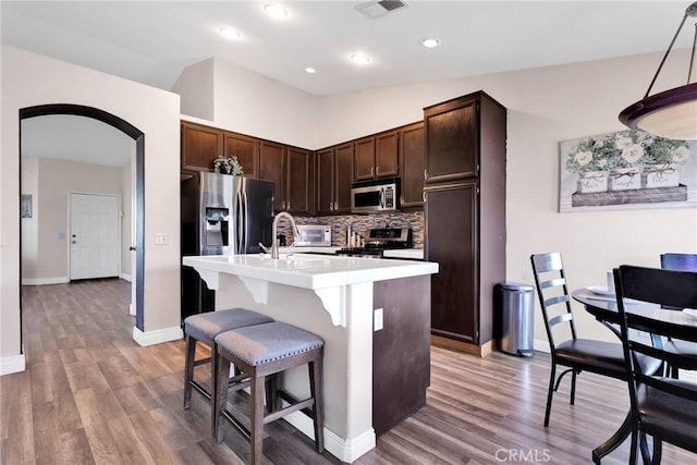 kitchen with arched walkways, dark brown cabinetry, appliances with stainless steel finishes, tasteful backsplash, and a center island with sink