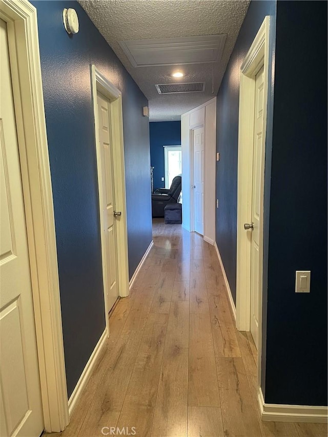 hall with a textured ceiling, light wood-type flooring, visible vents, and baseboards