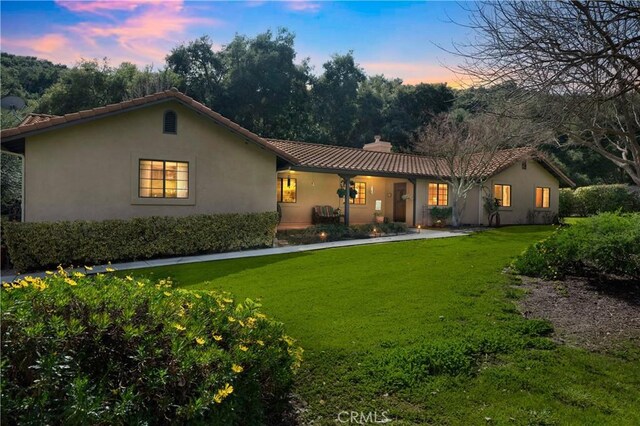 back of property at dusk featuring a tiled roof, a lawn, a chimney, and stucco siding