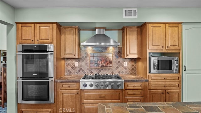 kitchen with visible vents, wall chimney range hood, appliances with stainless steel finishes, backsplash, and tile counters