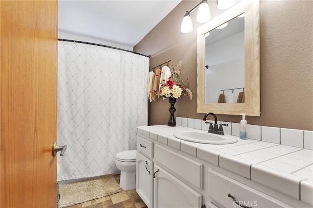 full bath with toilet, a textured wall, stone finish flooring, and vanity