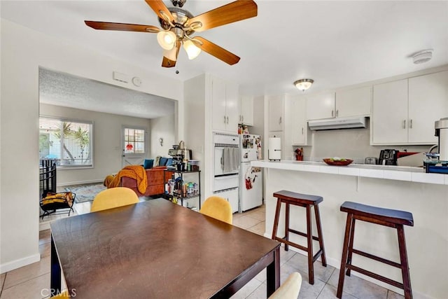 kitchen featuring tile countertops, under cabinet range hood, a kitchen bar, white cabinetry, and light tile patterned flooring