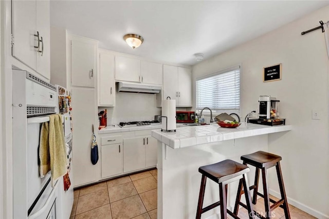 kitchen with under cabinet range hood, a peninsula, a sink, white cabinets, and tile counters