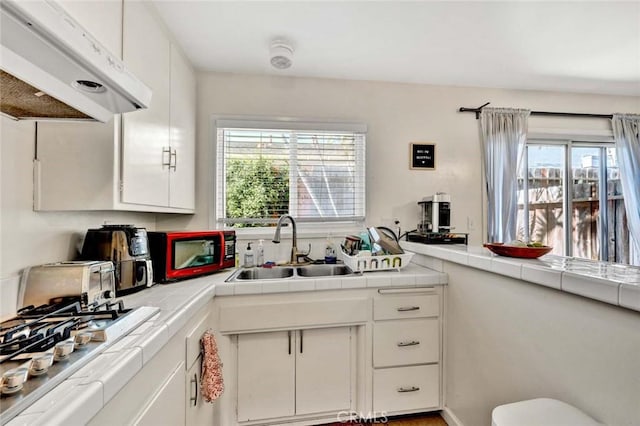 kitchen featuring tile countertops, a sink, white cabinets, and under cabinet range hood