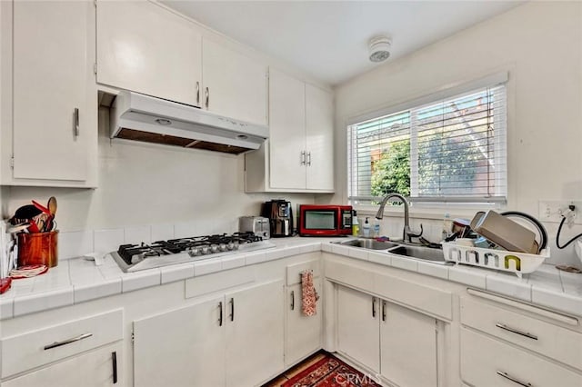 kitchen featuring tile countertops, under cabinet range hood, stainless steel gas stovetop, white cabinetry, and a sink