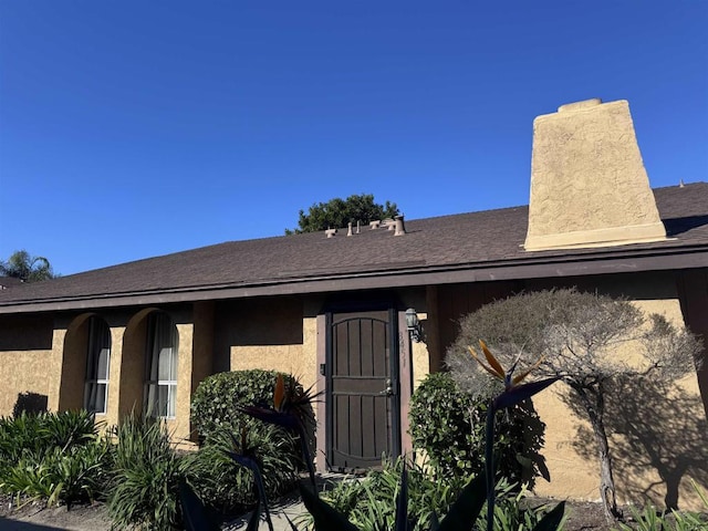 entrance to property featuring a shingled roof, a chimney, and stucco siding