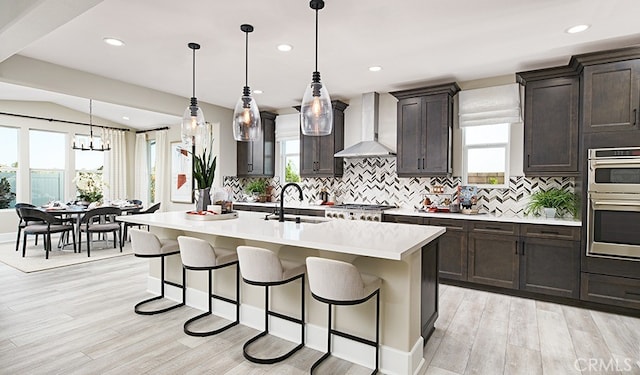 kitchen with a kitchen island with sink, light countertops, wall chimney range hood, pendant lighting, and a sink