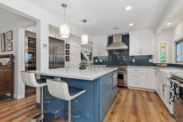 kitchen with high end appliances, hanging light fixtures, white cabinets, a kitchen island, and wall chimney range hood