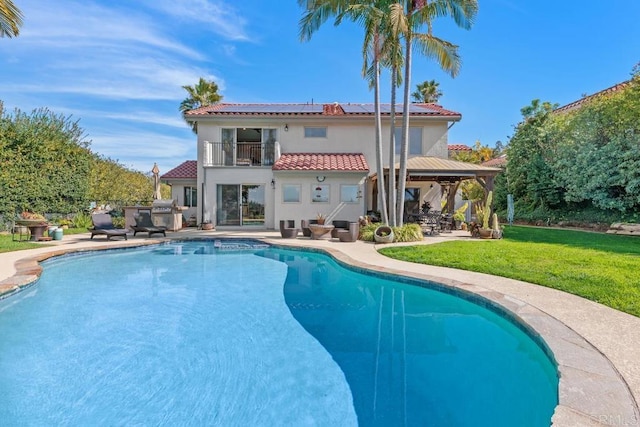 rear view of property with a tile roof, stucco siding, a lawn, roof mounted solar panels, and a balcony