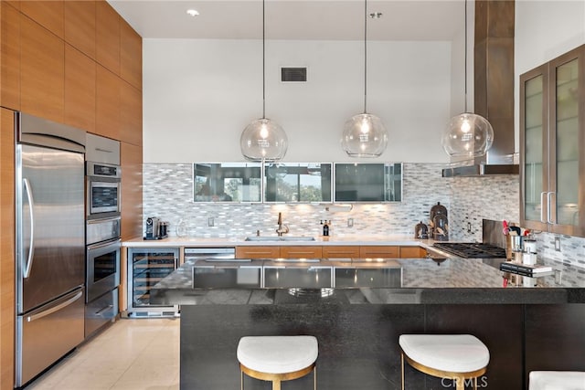 kitchen with a breakfast bar area, beverage cooler, appliances with stainless steel finishes, wall chimney range hood, and glass insert cabinets