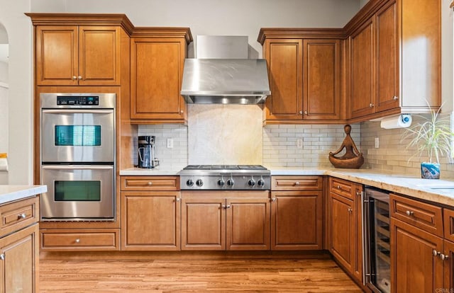 kitchen featuring wall chimney range hood, beverage cooler, stainless steel appliances, and brown cabinetry