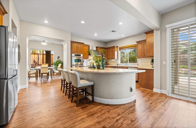 kitchen with arched walkways, brown cabinets, appliances with stainless steel finishes, a kitchen island, and light wood-type flooring