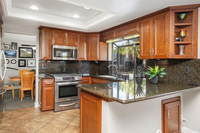 kitchen featuring a sink, appliances with stainless steel finishes, dark stone counters, a tray ceiling, and brown cabinetry