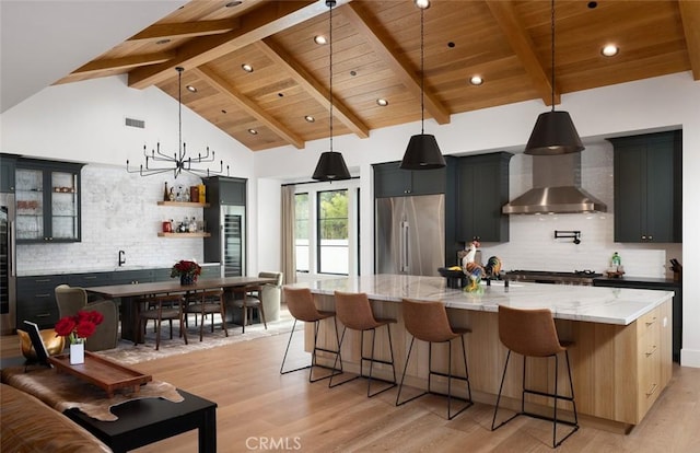 kitchen with built in fridge, a large island, hanging light fixtures, glass insert cabinets, and wall chimney exhaust hood