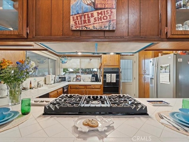 kitchen featuring glass insert cabinets, tile countertops, brown cabinetry, black appliances, and a sink