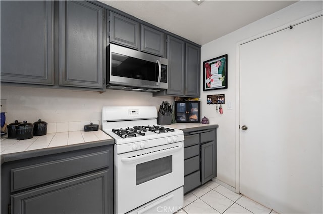 kitchen with tile countertops, stainless steel microwave, white gas range oven, and gray cabinets