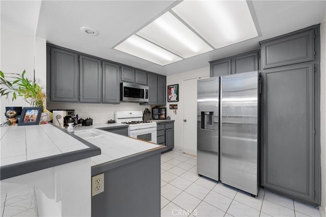 kitchen with stainless steel appliances, a peninsula, a sink, gray cabinets, and tile counters