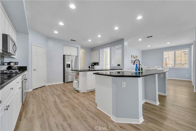 kitchen featuring appliances with stainless steel finishes, dark countertops, light wood-style flooring, and a kitchen island with sink