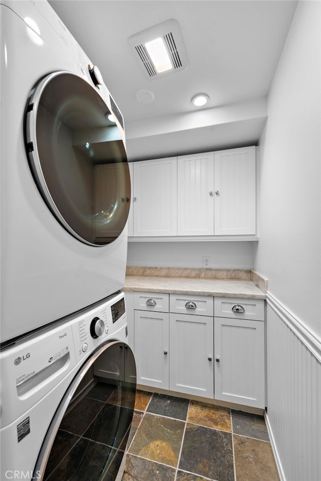 laundry area featuring visible vents, stone finish flooring, stacked washer and dryer, wainscoting, and cabinet space