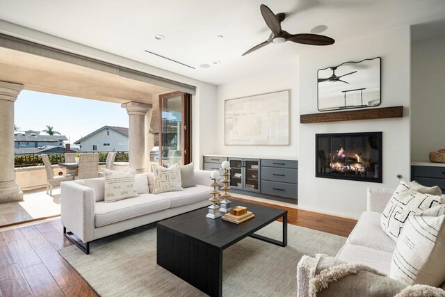 living area featuring a glass covered fireplace, ceiling fan, wood finished floors, a wall of windows, and ornate columns