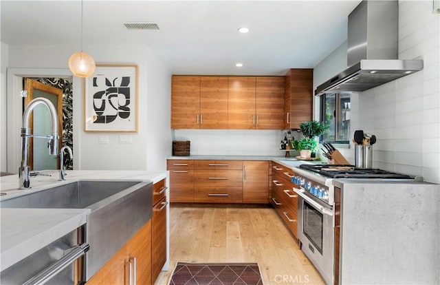 kitchen featuring stainless steel range, wall chimney range hood, brown cabinetry, and modern cabinets