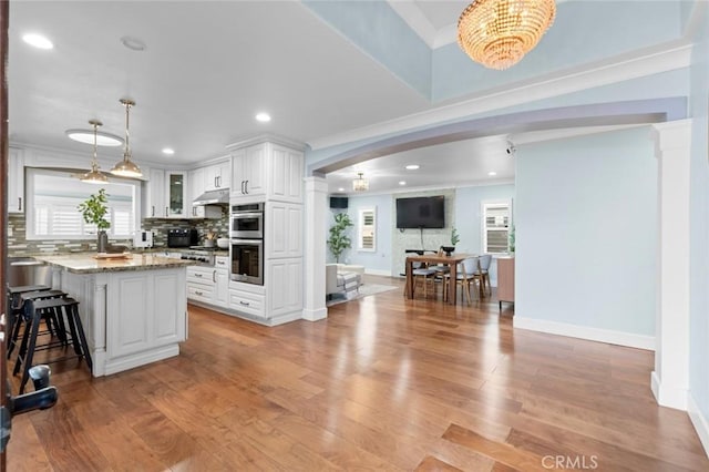 kitchen featuring double oven, under cabinet range hood, a healthy amount of sunlight, light wood-style floors, and tasteful backsplash