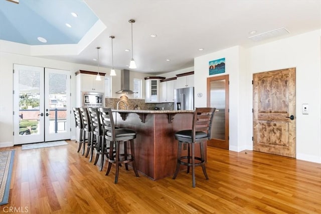 kitchen with stainless steel appliances, wall chimney range hood, french doors, light wood-type flooring, and a kitchen bar