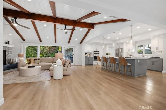 living area with a tiled fireplace, light wood-style flooring, ceiling fan, vaulted ceiling with beams, and recessed lighting