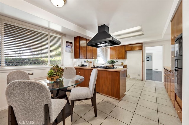 kitchen with light tile patterned floors, light countertops, black oven, brown cabinetry, and island exhaust hood